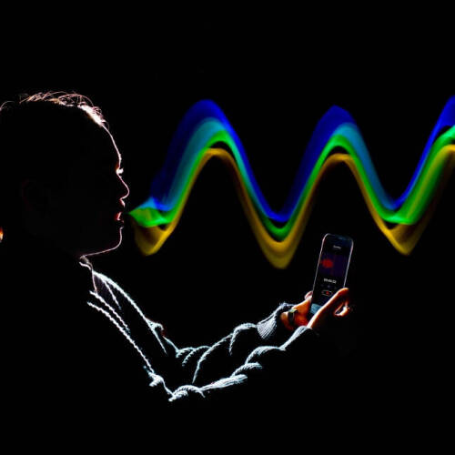 A silhouetted person holds a smartphone covered in rim lighting, appearing to record or speak into it while luminous green, yellow, and blue waves emanate in front of them, suggesting sound or data transmission.